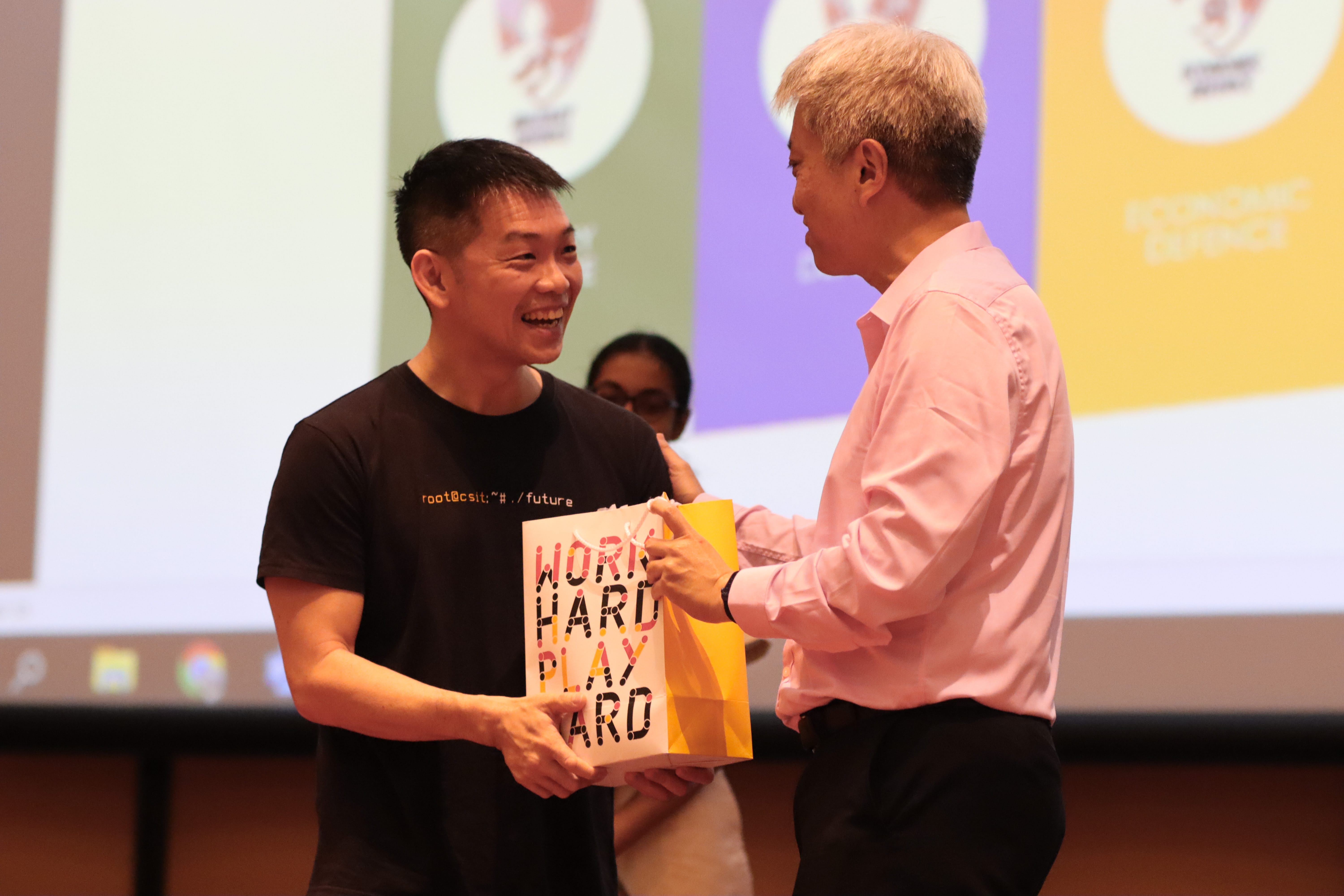 Two men exchange a gift bag reading "Work Hard Play Hard" on a stage, one smiling in a black shirt.